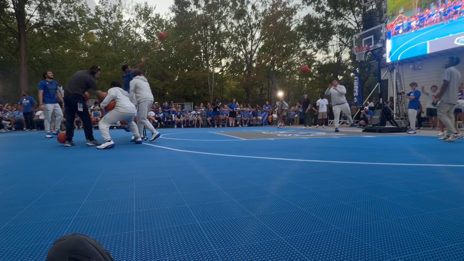 Participants shooting basketballs on the blue outdoor court as spectators watch from the sideline